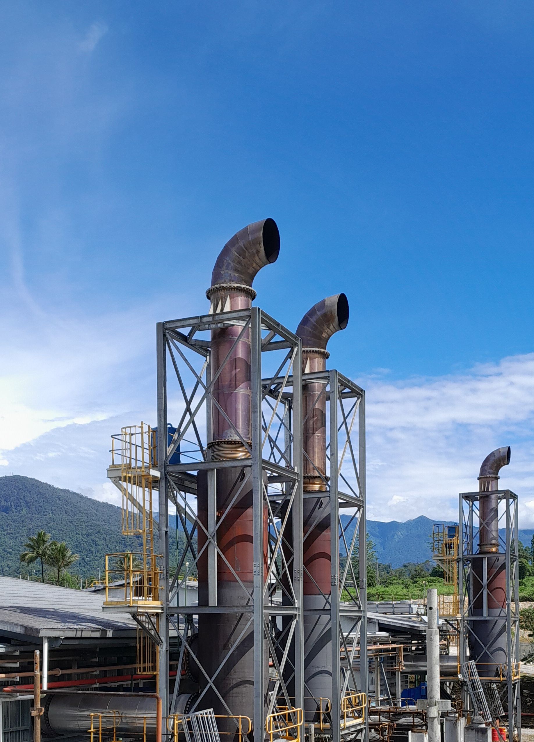Outdoor industrial smokestacks at a power plant against a clear blue sky with mountains